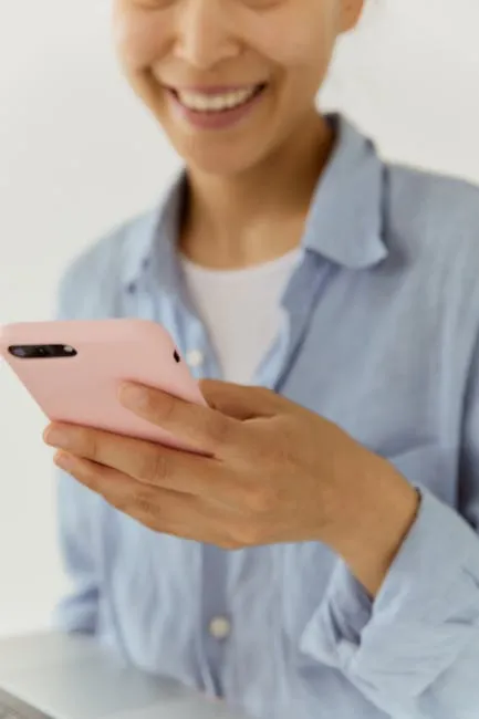 Close-up of a woman smiling while holding a pink smartphone indoors. Casual and friendly atmosphere.